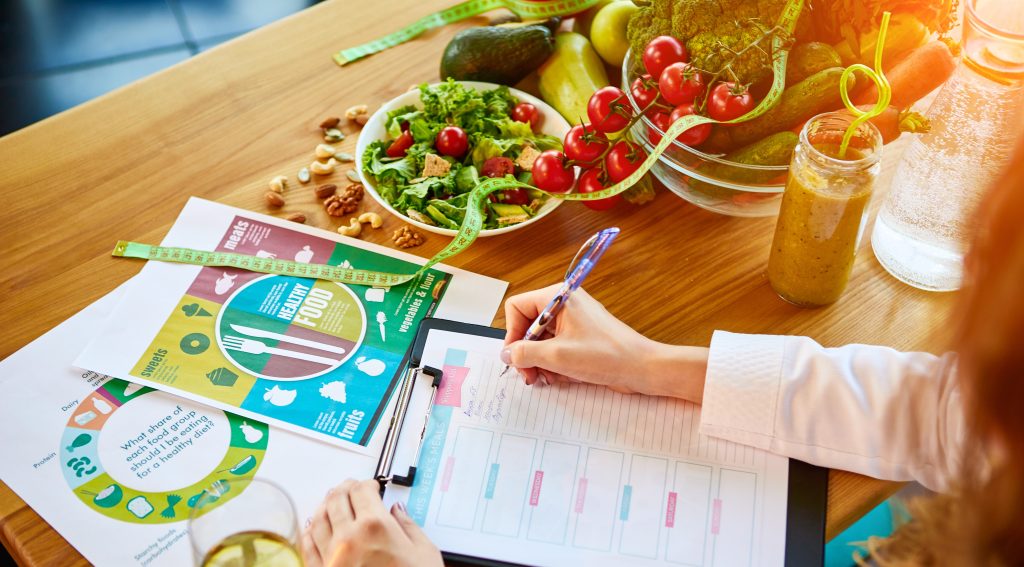 Woman dietitian in medical uniform with tape measure working on a diet plan sitting with different healthy food ingredients.