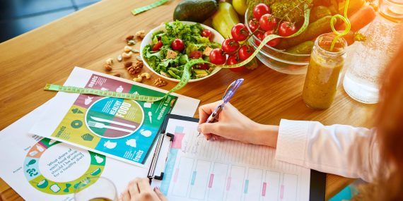 Woman dietitian in medical uniform with tape measure working on a diet plan sitting with different healthy food ingredients.