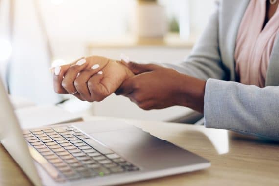 Woman, laptop and hands with wrist pain, injury or carpal tunnel syndrome by office desk.