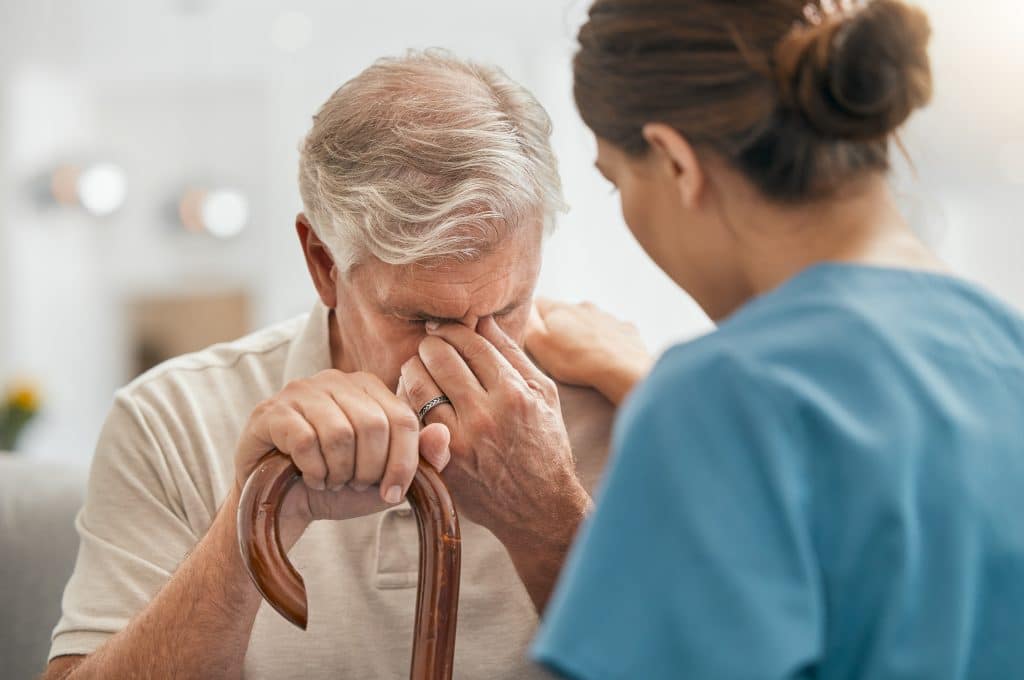 Chiropractor speaking with a tired, elderly male patient. The patient is holding a walking cane in one hand and pinching the space between his eyes with his other hand.